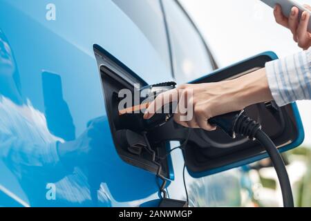 Transportation. Young woman on electric car having stop at charging station standing plugging charger using app on smartphone while charging close-up Stock Photo