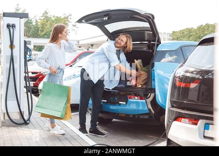 Traveling. Young couple traveling by electric car stop at charging station putting shopping bags into trunk looking at each other smiling cheerful Stock Photo