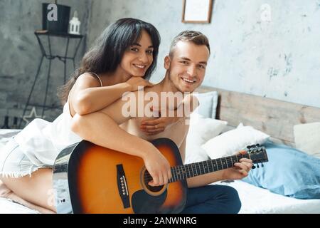 Mixed Race Couple. Young man playing guitar while woman hugging him sitting on bed at home looking camera cheerful Stock Photo