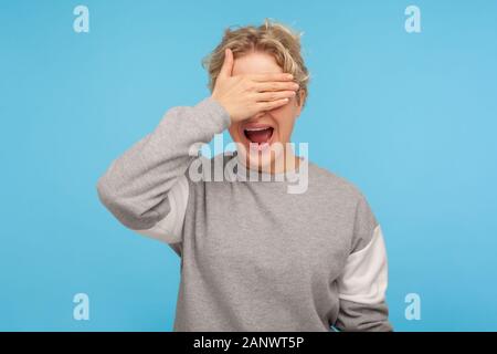 Don't want to look at this. Happy woman with curly hair in sweatshirt covering eyes and screaming with wide opened mouth, scared or ashamed to watch. Stock Photo