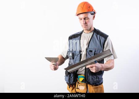 The builder is cleaning a spatula. In work clothes and hard hat. On a light gray background. Stock Photo