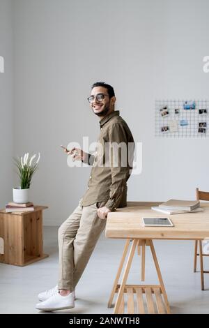 Young cheerful businessman using smartphone while standing by table Stock Photo