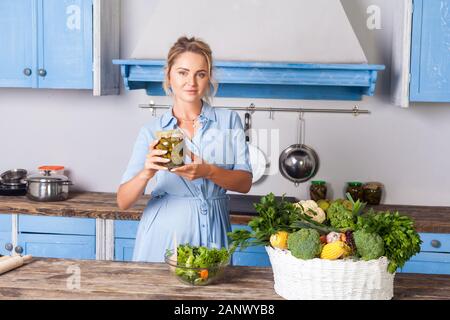 Young pretty woman holding jar of pickles and smiling looking at camera, cooking green salad in modern kitchen, preparing vegetarian meal, basket of f
