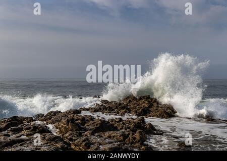 Wave breaking on rocky shoreline at Leo Carrillo State Beach, California. Rocks in foreground; pacific ocean, cloudy blue sky in background. Stock Photo