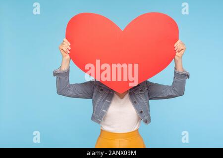 Anonymous woman in casual outfit hiding face behind huge red heart, concept of secret love fondness, unknown lover and affair, romance on Valentines d Stock Photo