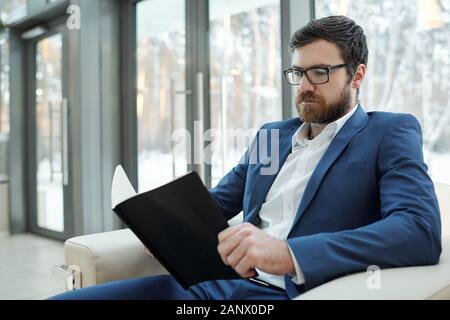 Serious young bearded man in formalwear and eyeglasses looking through notes Stock Photo