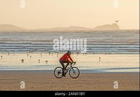 Pembrey, Wales. 19th Jan 2020. UK Weather: A lone road cyclist rides his bike across the sand at Cefn Sidan Beach near Llanelli in Carmarthenshire late this afternoon with the iconic Worms Headland on the Gower Peninsula looms large in the distance. Credit: Phil Rees/Alamy Live News