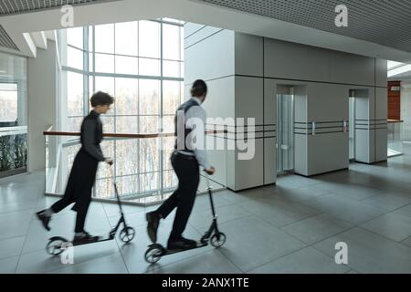 Blurred outlines of young managers moving on scooters in corridor Stock Photo