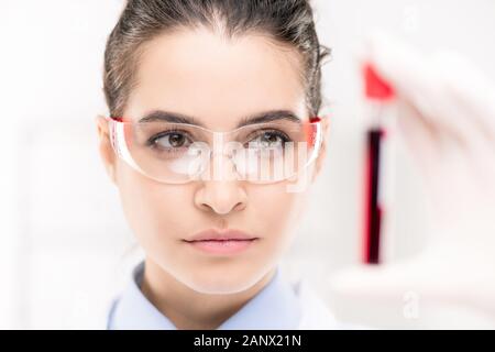 Pretty young serious female looking through protective eyeglasses at flask Stock Photo
