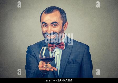 A mature happy man holding showing offering a glass of red wine looking at you camera smiling happily. Mixed race bearded model isolated gray backgrou Stock Photo