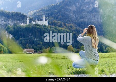 Young blond haired girl squatting backwards on a green field, holding a photo camera and taking pictures from far away of the Neuschwanstein castle in Stock Photo