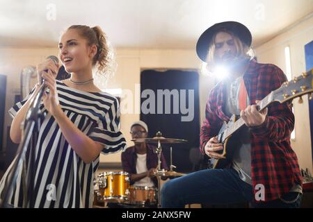 Portrait of smiling young woman singing to microphone during rehearsal or concert with music band Stock Photo