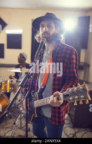 Waist up portrait of contemporary long-haired man playing guitar and singing to microphone during rehearsal or concert with music band Stock Photo