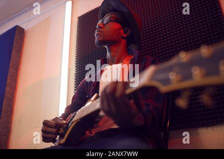 Low angle portrait of contemporary African-American man playing guitar sitting on high chair while writing music in dimly lit studio, copy space Stock Photo