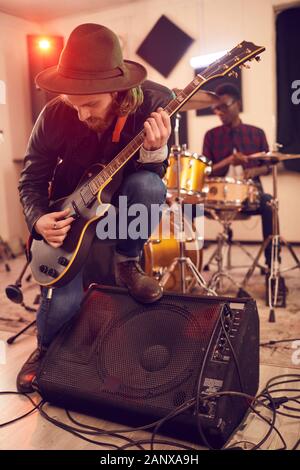 Full length portrait of contemporary young man playing electric guitar solo leaning on amplifier during concert or rehearsal with music band in background Stock Photo