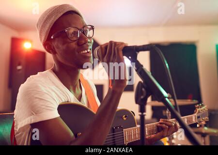 Portrait of contemporary African-American man singing to microphone and playing guitar during rehearsal or concert with music band in recording studio, copy space Stock Photo