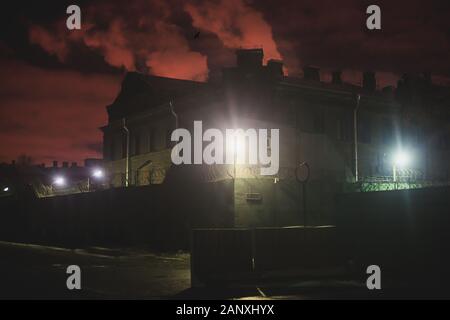 A night view of protected creepy penitentiary prison building facility, asylum exterior with barded wire on walls, red night sky, concept of scary hou Stock Photo