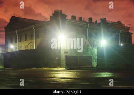 A night view of protected creepy penitentiary prison building facility, asylum exterior with barded wire on walls, red night sky, concept of scary hou Stock Photo