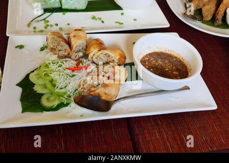 Fried spring rolls with with sliced cucumber and sweet chili sauce in a white plate on red table, Vietnamese food in Cambodia Stock Photo