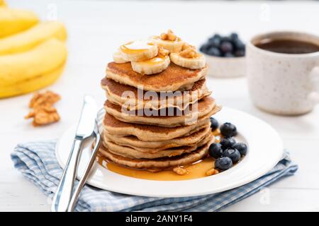 Tasty pancakes with banana and syrup served with fresh blueberries and cup of black coffee on a white table. Closeup view sweet breakfast food