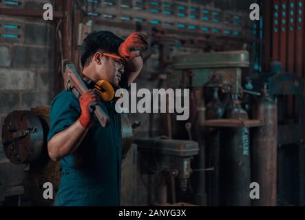 Asian male engineer in repair garage wiping sweat away from his head while working during long day shift and holding industrial hand tools Stock Photo