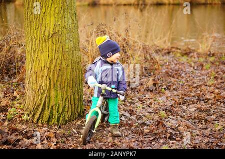 POZNAN, POLAND - Jan 12, 2020: Young boy sitting on a small running bike next to a tree by a lake in the Debiec forest. Stock Photo