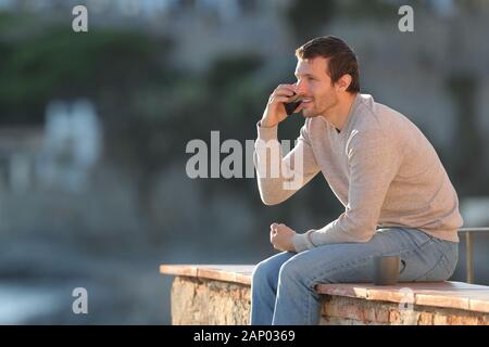 Happy man calls on mobile phone sitting on a wall in a coast town on vacations Stock Photo