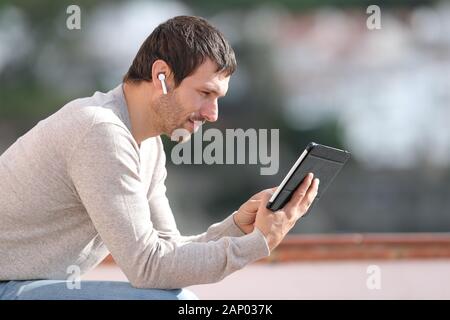 Serious man watching media with tablet and wireless earbuds sitting in a rural town Stock Photo