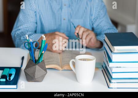 Close-up hands of the teacher man is preparing for the lesson: reading a book and making notes at the table, next to a pile of books and stationery, e Stock Photo