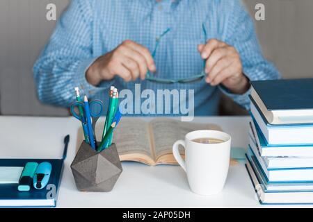 Close-up hands of the teacher man is preparing for the lesson: reading a book and making notes at the table, next to a pile of books and stationery, e Stock Photo