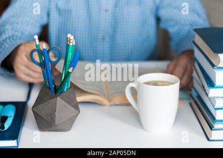 Close-up hands of the teacher man is preparing for the lesson: reading a book and making notes at the table, next to a pile of books and stationery, e Stock Photo