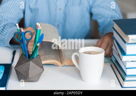 Close-up hands of the teacher man is preparing for the lesson: reading a book and making notes at the table, next to a pile of books and stationery, e Stock Photo
