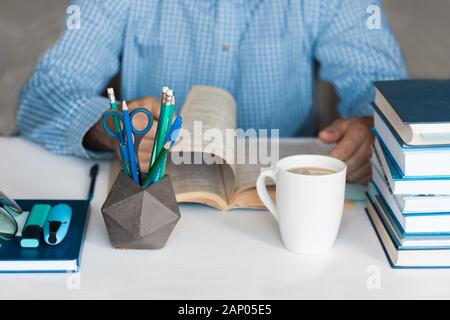 Close-up hands of the teacher man is preparing for the lesson: reading a book and making notes at the table, next to a pile of books and stationery, e Stock Photo
