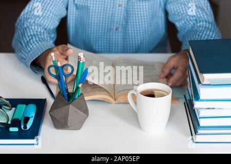 Close-up hands of the teacher man is preparing for the lesson: reading a book and making notes at the table, next to a pile of books and stationery, e Stock Photo