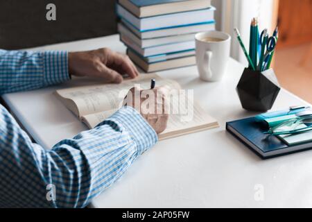 Close-up hands of the teacher man is preparing for the lesson: reading a book and making notes at the table, next to a pile of books and stationery, e Stock Photo
