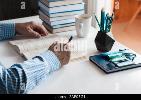 Close-up hands of the teacher man is preparing for the lesson: reading a book and making notes at the table, next to a pile of books and stationery, e Stock Photo