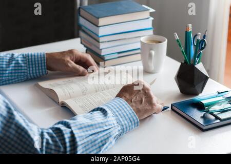 Close-up hands of the teacher man is preparing for the lesson: reading a book and making notes at the table, next to a pile of books and stationery, e Stock Photo