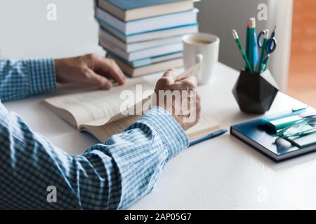 Close-up hands of the teacher man is preparing for the lesson: reading a book and making notes at the table, next to a pile of books and stationery, e Stock Photo