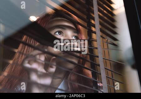 Asian woman looking through window blinds spying on neighbours - Young lonely millennial woman peeping through glass observing gossip and action outdoors - introvert, spy and intrusive concepts