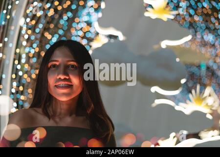 Young diverse woman smiling alone at night surrounded by colourful neon bokeh lights - Cute hipster American Asian girl with orange glow on face in club - Nightlife, dinner date and lifestyle concept Stock Photo