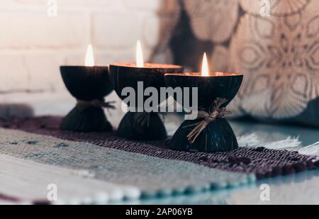 Burning spa aroma candle in coconut shell on a knitted multicolored rug and gray curtains opposite the white wall, cozy home interior Stock Photo