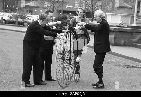 1980s, a young woman attempting to get on a penny-farthing bicycle, being held steady by some male helpers. Also known as the 'high wheeler' - as can be seen why in the picture - the machine was the first to be called a 'bicycle'. The 'ordinary' as it was also known as, had a large front wheel providing speed and comfort but became obsolete with the new 'safety' bicycles from the 1880s, which provided similar speed, comfort from their pneumatic tyres and with a reduced danger due far lower height to fall from. The name came from the British ''penny' and 'farthing' coins and symbolic of its era Stock Photo