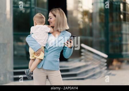 Successful business woman in blue suit with baby. Successful mother in business walking with baby in her hands and texting message on cellphone Stock Photo
