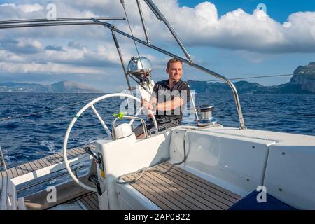 Sailor at the helm of modern sailing yacht. Mediterranean sea, near Ischia island and Capri island on the background, Italy. Stock Photo