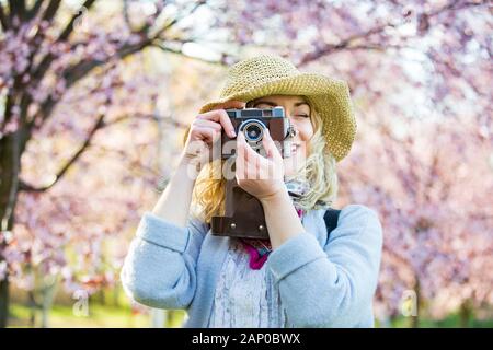 Portrait of beautiful woman in straw hat traveling in beautiful park with cherry trees in bloom, taking photos on retro camera. Tourist with backpack Stock Photo