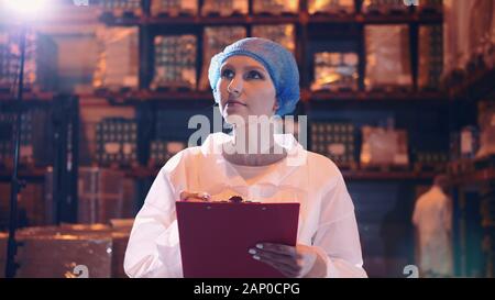 Controller checking factory storage room. Pretty young lady walking between shelves in huge storage room and making notes during inspection in factory. Stock Photo