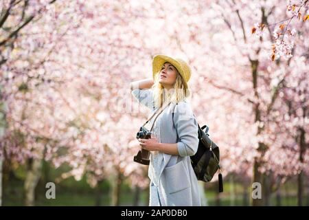Portrait of beautiful woman in straw hat traveling in beautiful park with cherry trees in bloom, taking photos on retro camera. Tourist with backpack Stock Photo