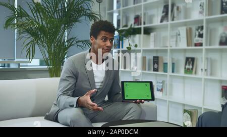 Young African-American man in suit sitting in office on sofa with anonymous colleague and making presentation with tablet with tablet with green chromakey screen. Stock Photo