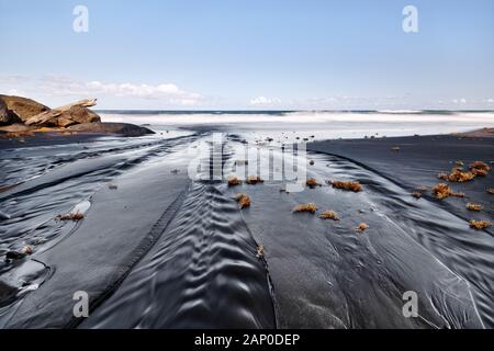 Beach with dark sand and some rocks, in the foreground sand ribs, which were formed by a small river, some tufts of tufts lie on the sand, shot from d