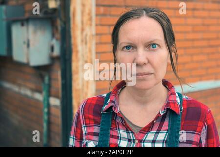 Serious female farmer posing on farm. Confident woman farm worker wearing plaid shirt and jeans overalls looking at camera. Stock Photo